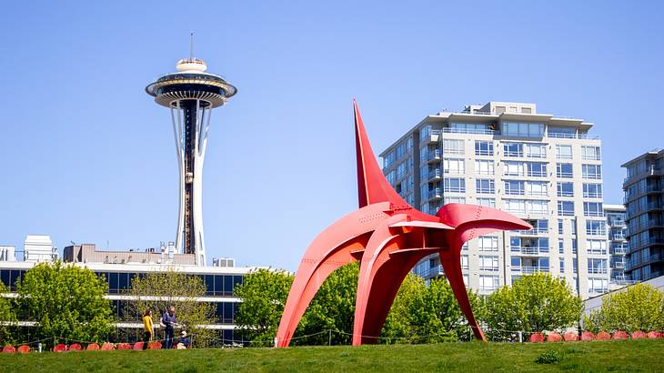 A red sculpture on the grass next to trees, buildings, and an observation tower