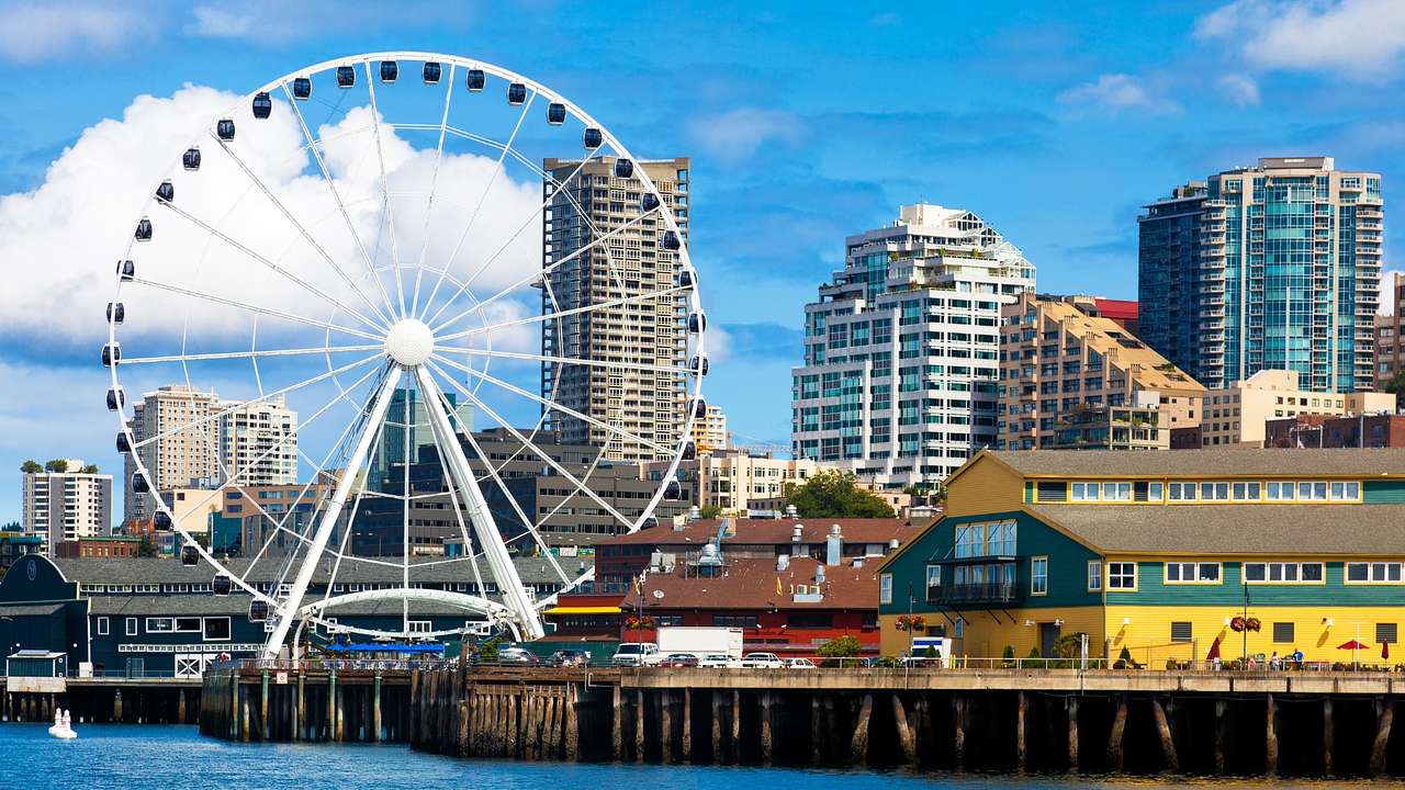 A Ferris wheel next to buildings, a pier, and a body of water under a blue sky