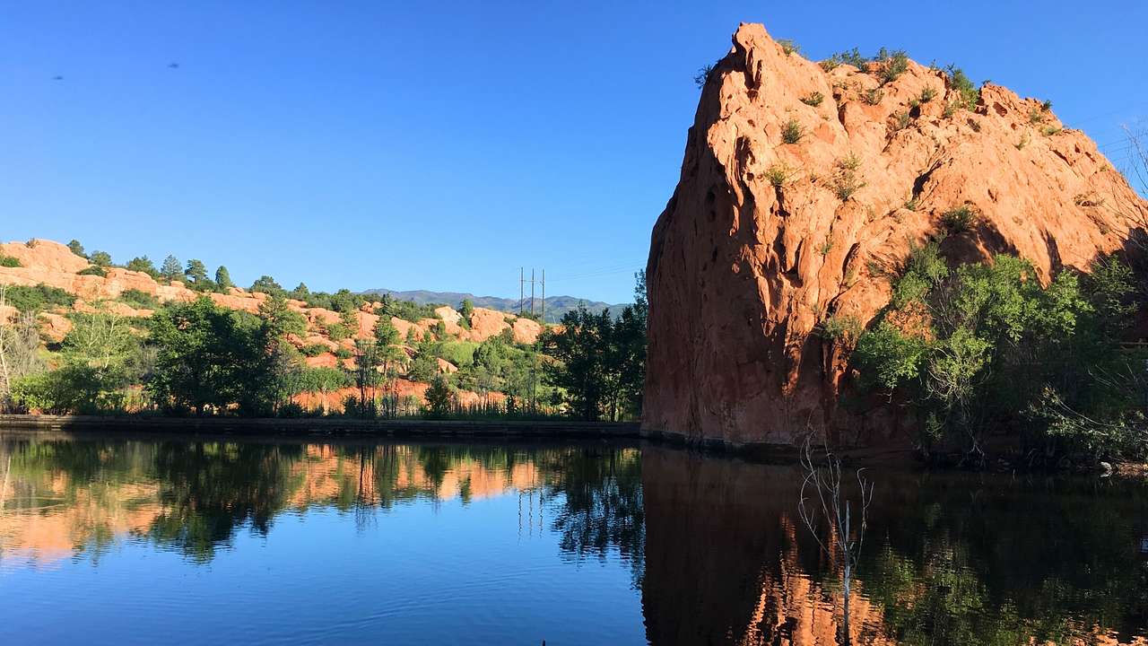 Red rock mountains with greenery on them next to a pond under a blue sky