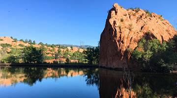 Red rock mountains with greenery on them next to a pond under a blue sky
