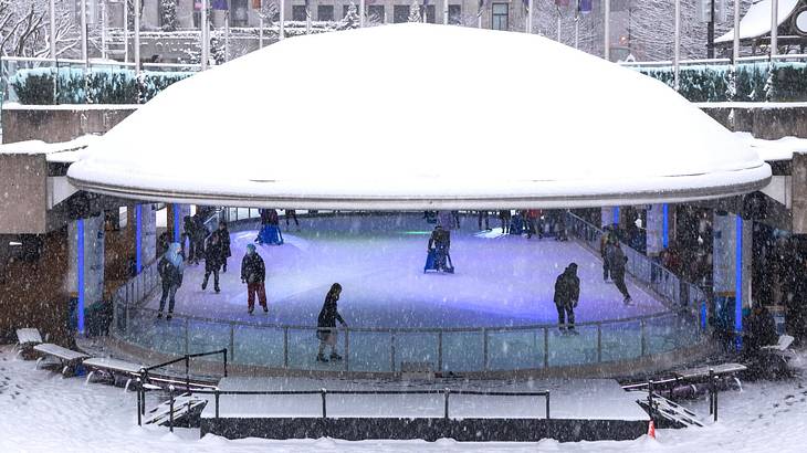 A view looking into an ice rink with people skating as it snows