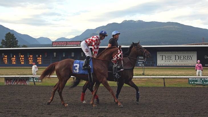 Two jockeys riding horses on a race track with mountains behind it