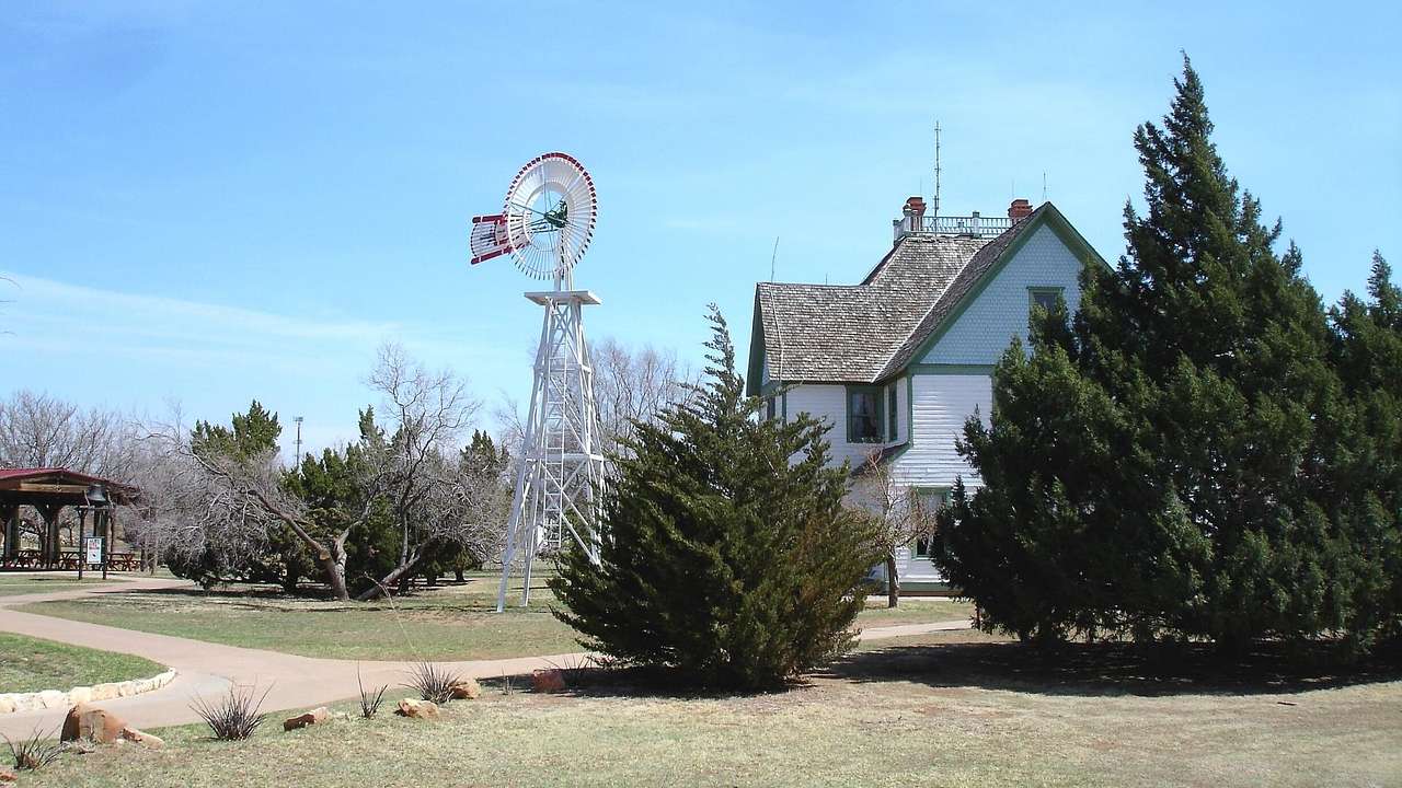 An American windmill next to a small house, grass, and trees under a blue sky