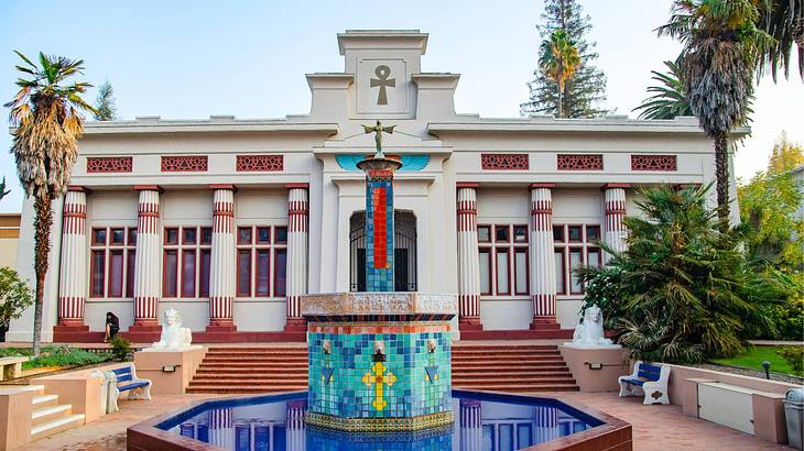 A stone building with columns next to palm trees and a water fountain with a mosaic