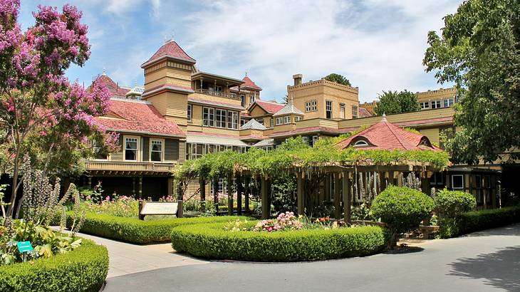 A mansion next to a garden with green bushes, pink flowers, and a pergola