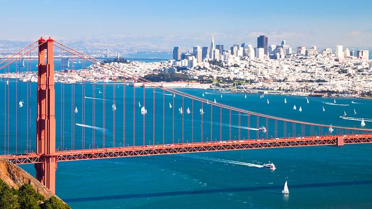 A red suspension bridge over a bay with a city in the background