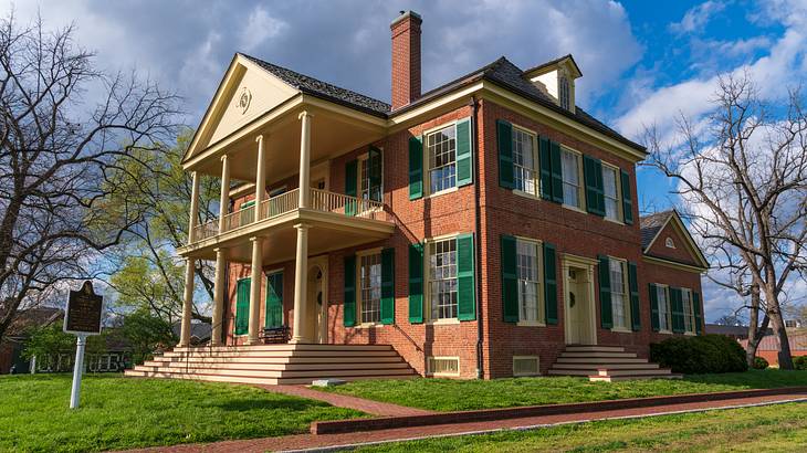 A red-brick mansion with columns and green window shutters and grass surrounding it