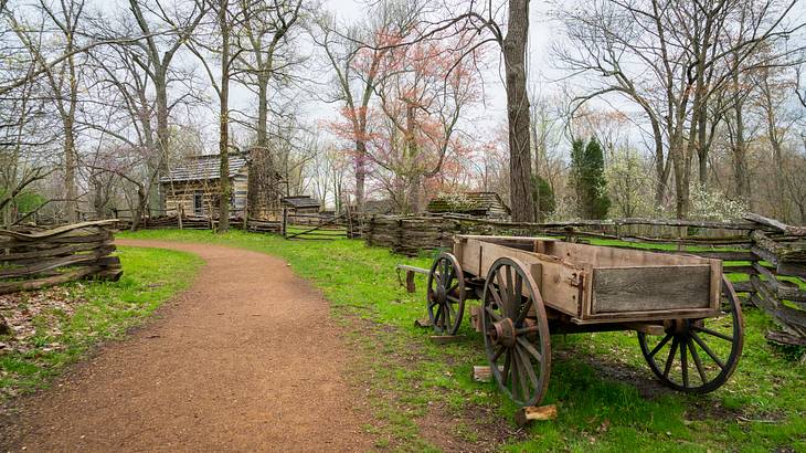 An old cart on green grass beside a pathway leading to an old hut-shaped house