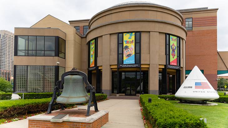 A contemporary round brown building with a huge bell at the entrance