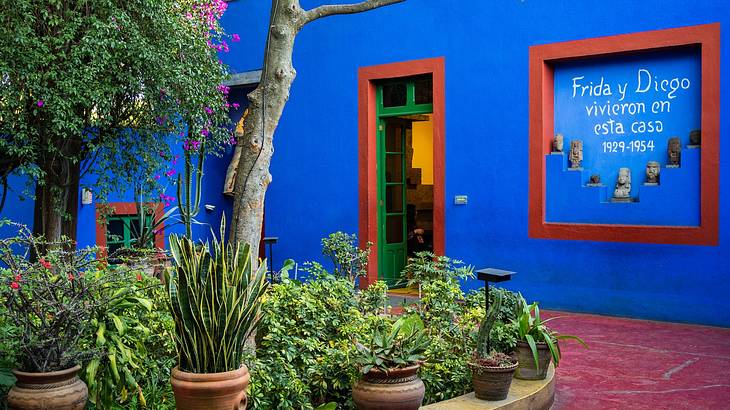 A blue building with a sign and a red door frame, next to green plants and a red path