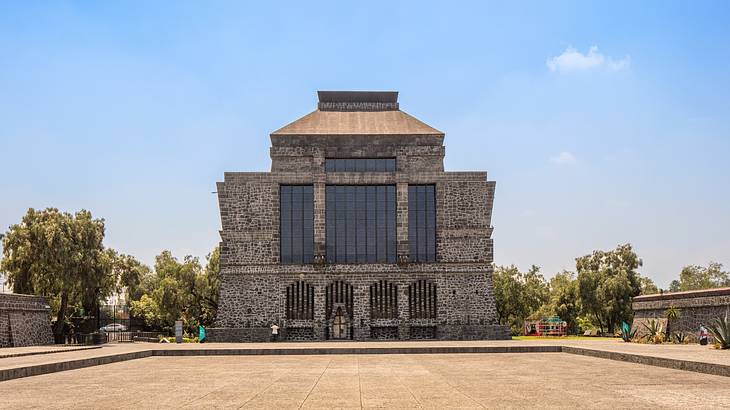 A stone temple-style structure with glass windows, next to a courtyard and trees