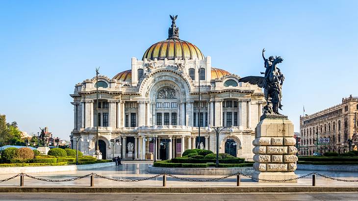 A white stone building with columns and a dome, next to a statue under a blue sky