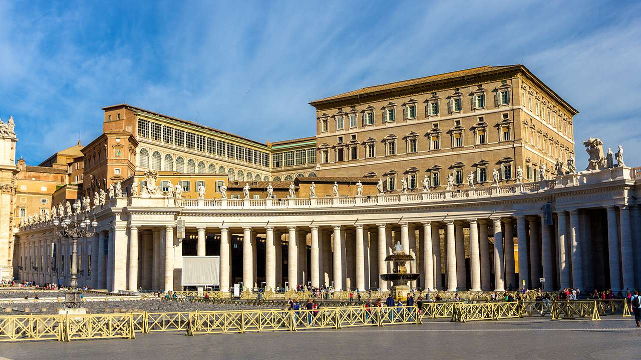 A palace with a row of columns featuring sculptures on top, facing a small fountain