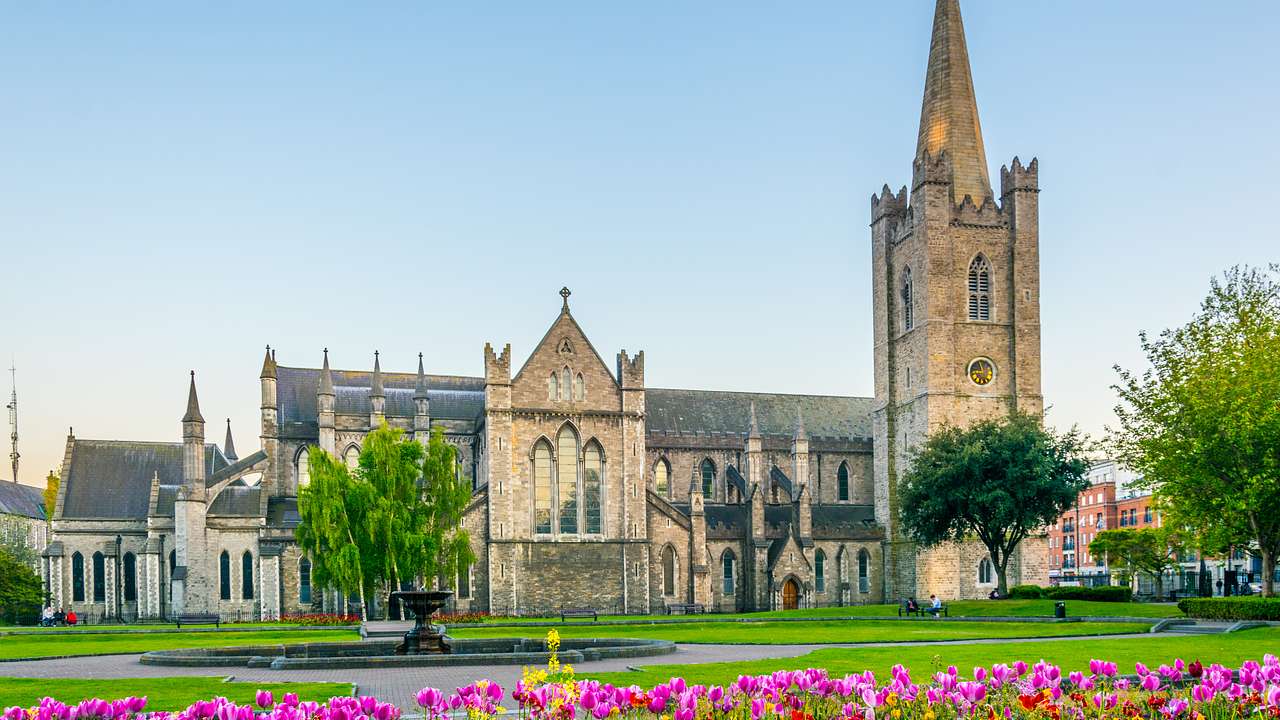 A stone cathedral with spires, a few trees, a fountain, and pink flowers in front