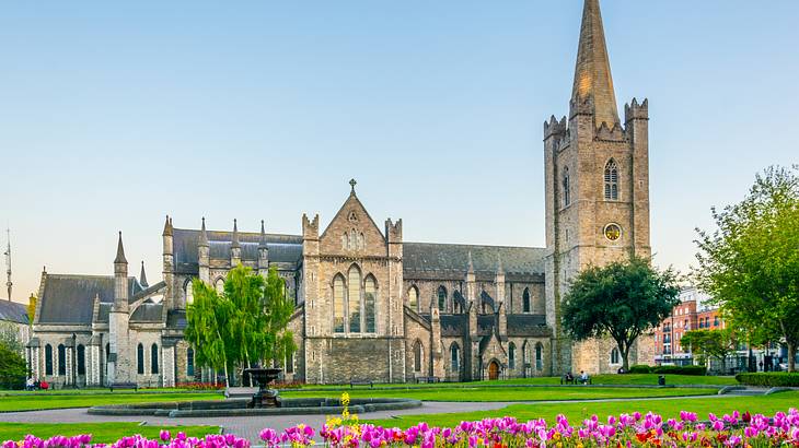 A stone cathedral with spires, a few trees, a fountain, and pink flowers in front