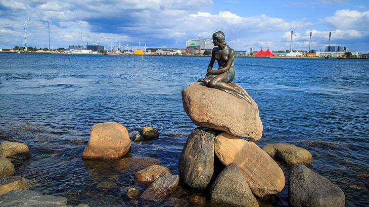 A bronze little mermaid statue on a tall pile of rocks above water