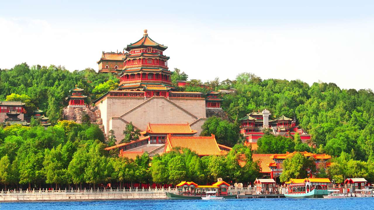A pagoda on top of a mountain, surrounded by pavilions and trees by a lake with boats
