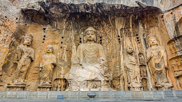 Large Buddhist statues carved out of a rock wall, behind a small barrier and a bowl