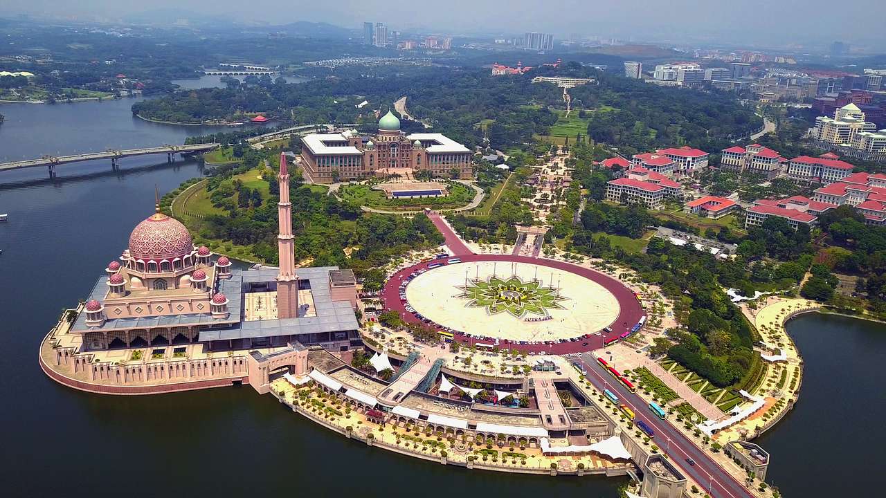 An aerial view of a vast, pink-colored mosque and greenery next to a lake
