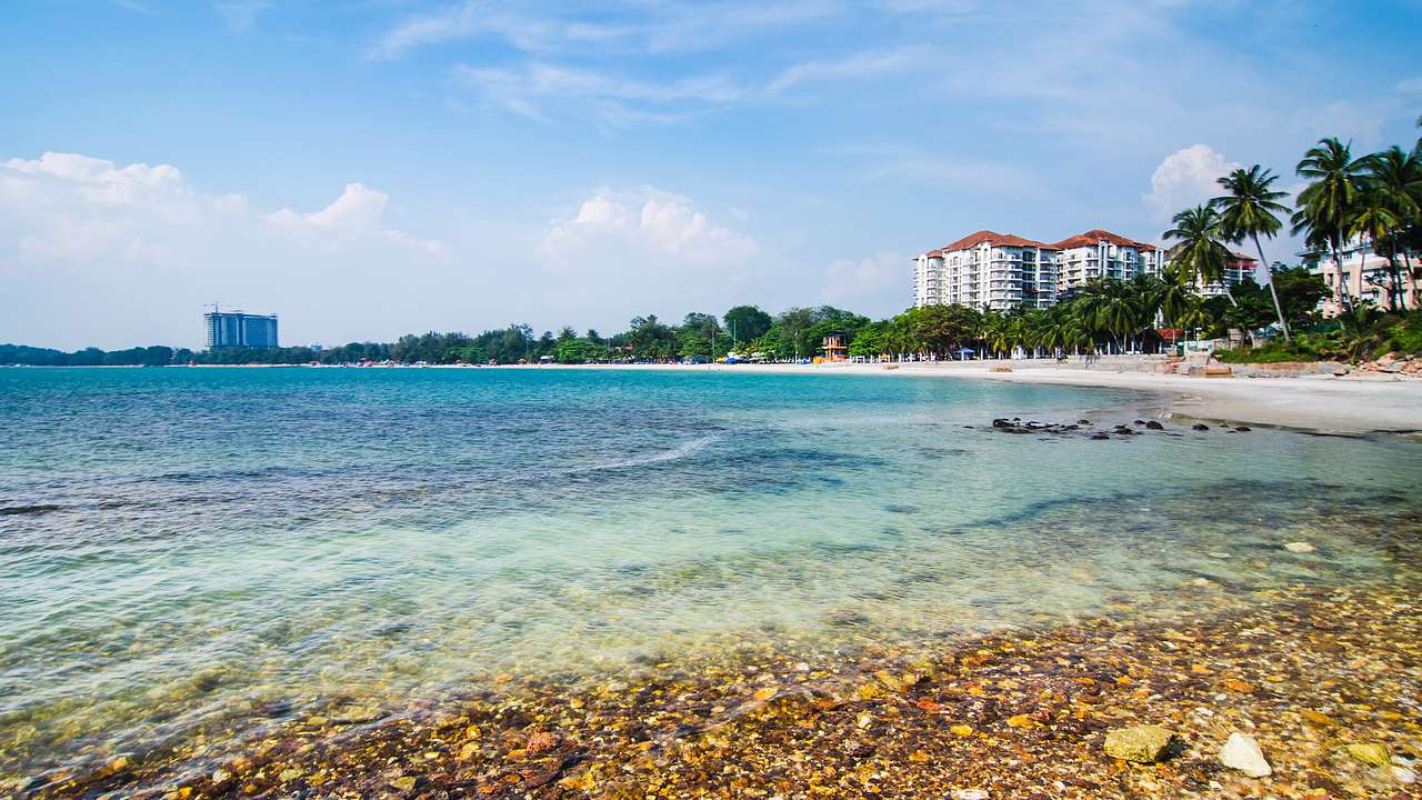 A pebble seashore next to crystal-clear water, buildings, and palm trees