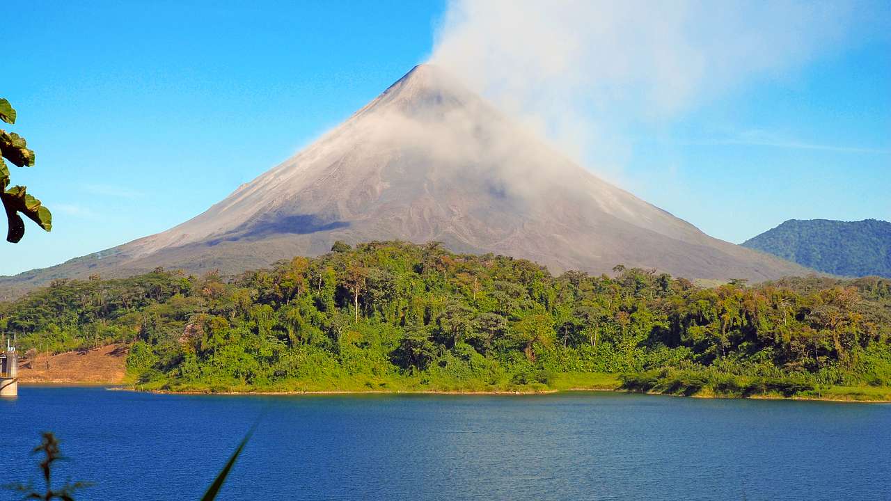 A volcano next to greenery and a body of water under a blue sky