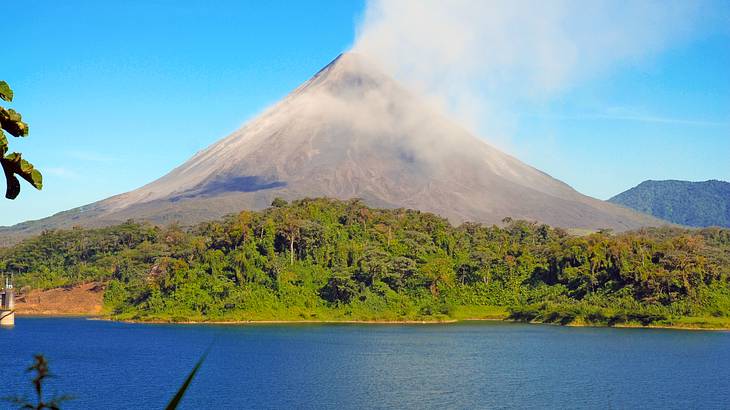 A volcano next to greenery and a body of water under a blue sky