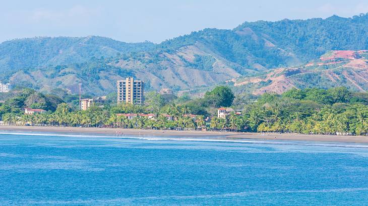 Blue sea with buildings, shoreline, palm trees, and mountains in the background