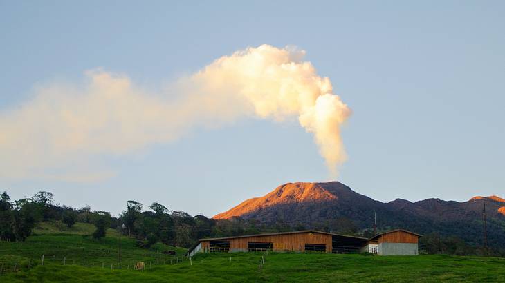 A house on a grass-covered land with the volcano giving off a white smoke in the back