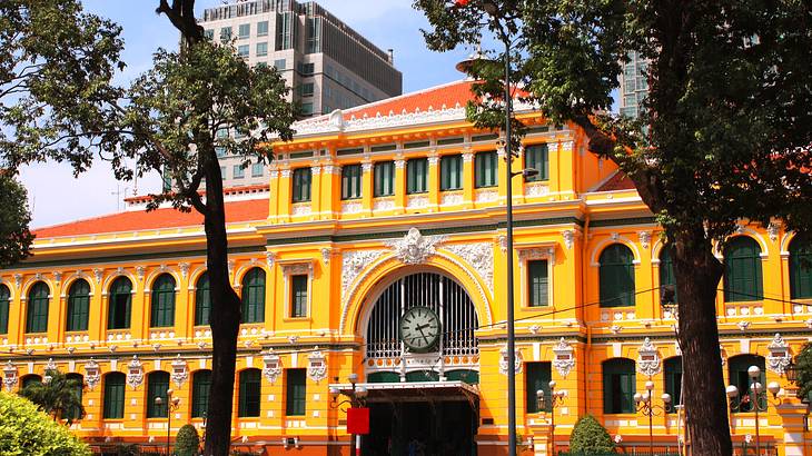 A yellow and orange building with a clock, trees in front, and a tall building behind