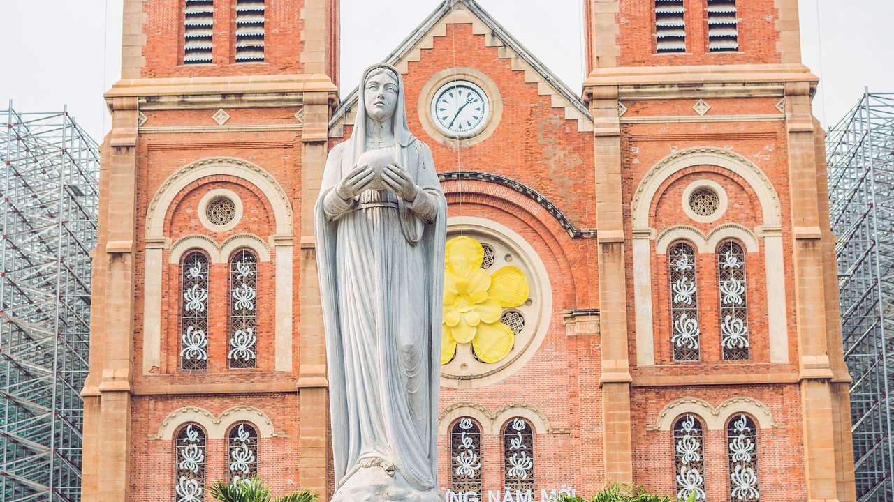 A stone statue of a woman in front of a red brick cathedral with a clock