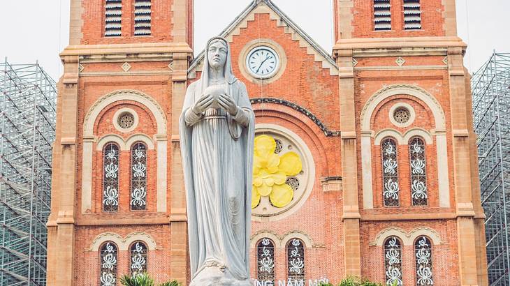 A stone statue of a woman in front of a red brick cathedral with a clock