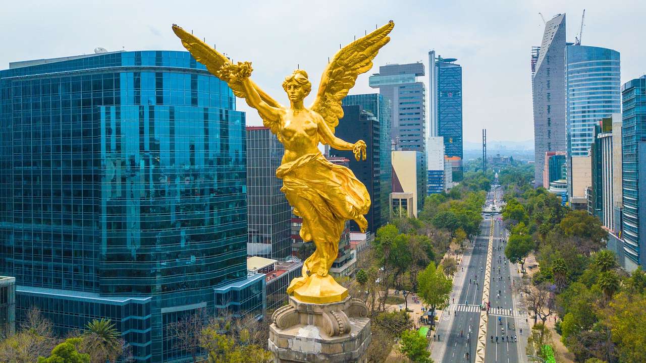A gold angel statue on top of a column, next to city buildings and a road below