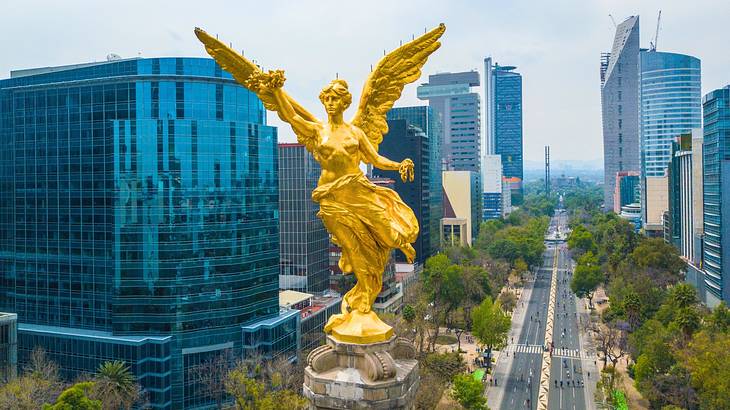 A gold angel statue on top of a column, next to city buildings and a road below