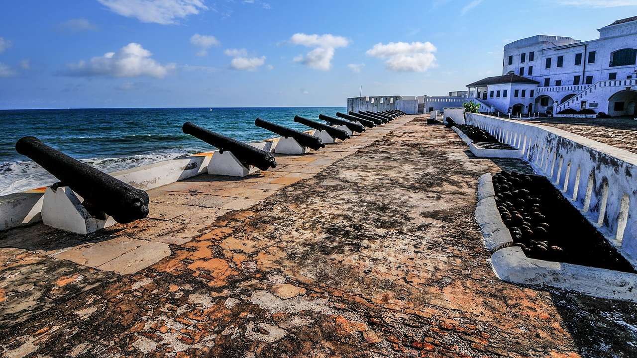 A walkway with cannons and ocean water to its left and a white building to its right
