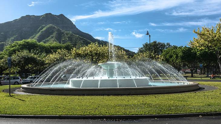 A fountain in a park with a mountain, and a 
partly cloudy sky in the background