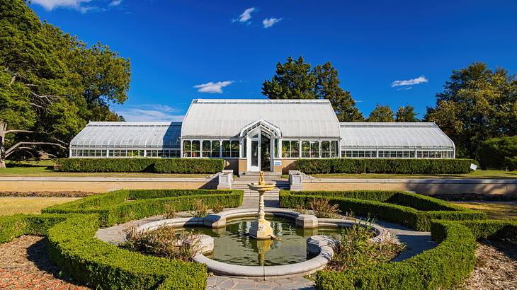 A landscaped garden with a fountain and hedges in front of a greenhouse