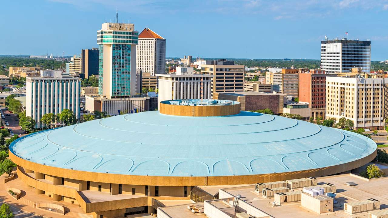 A large round building with a blue roof next to city buildings on a sunny day