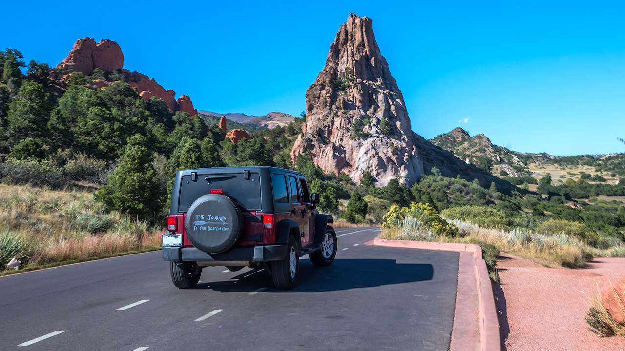 The back of a red and black Jeep on a road with greenery and a tall rock formation