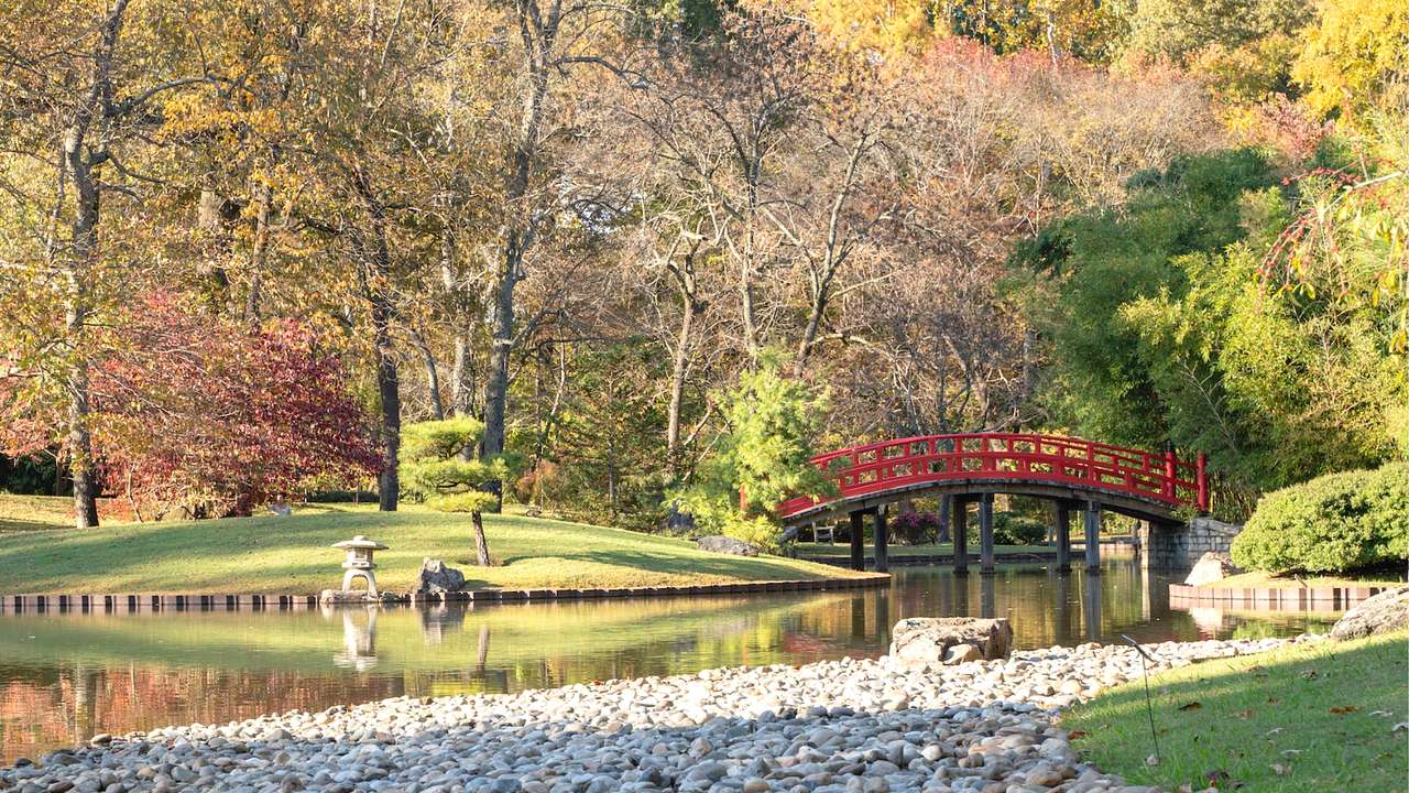 A small red bridge over a pond surrounded by grass, pebbles, and fall trees