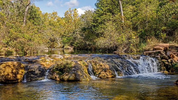 A small waterfall over rocks, against green trees under a partly cloudy sky