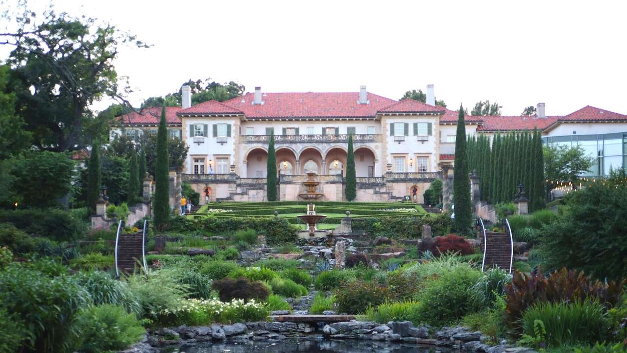 A fountain surrounded by a garden against a white mansion with a red roof