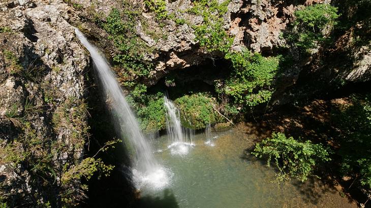 A rocky cliff with patches of bushes and a waterfall flowing down into a pool