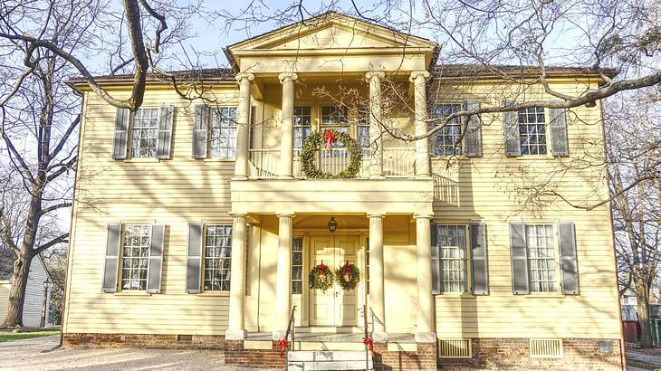 A yellow mansion-style house with wreaths on the door and bare trees surrounding