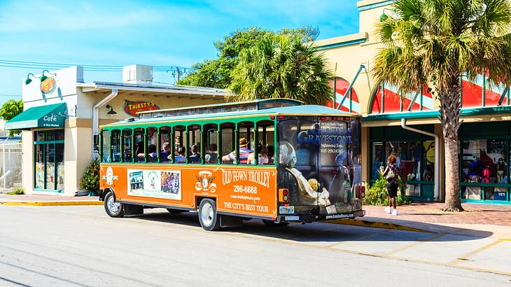An orange and green trolley vehicle on the street with buildings and palms behind it