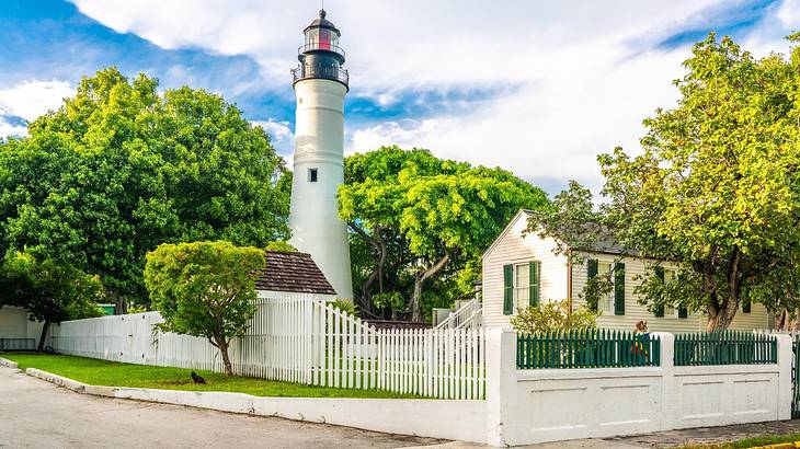 A white lighthouse next to a small house surrounded by greenery and a white fence