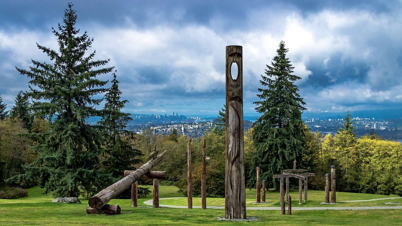 Totem poles on the grass surrounded by pine trees with a view of a city in the back