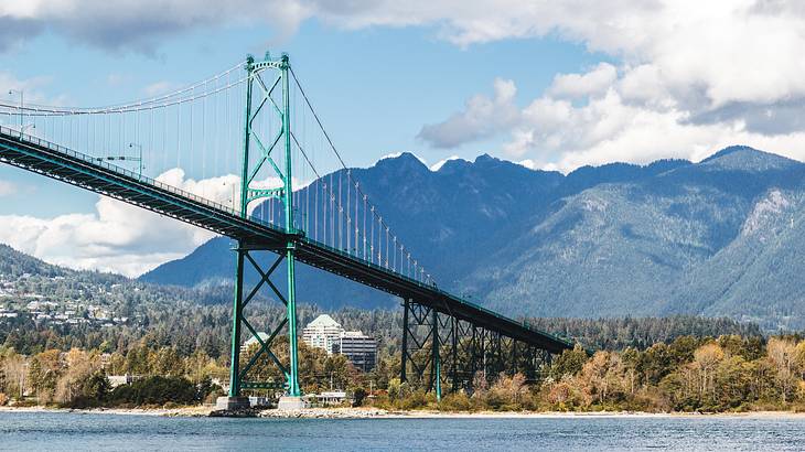 A green steel suspension bridge over a river with trees, buildings, and mountains