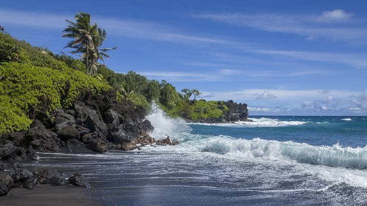 A black sand beach with waves crashing into the shore next to rocks with greenery