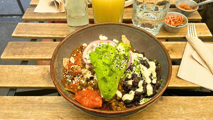 A bowl of food with beans, red sauce, guacamole and more on a wooden table