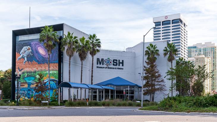 A white museum with a "Mosh" sign and a colorful mural next to palm trees